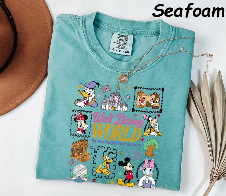 K&ouml;nnte beinhalten: T&uuml;rkisfarbenes T-Shirt mit einem farbenfrohen Grafikdesign von Disney-Figuren und dem Text "Walt Disney World". Das Design umfasst Donald Duck, Minnie Mouse, Chip und Chap, Goofy, Mickey Mouse und Daisy Duck. Das Shirt ist von der Marke Comfort Colors.