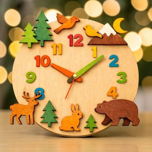 Op de afbeelding: Houten wandklok met een licht houten wijzerplaat, met kleurrijke cijfers en accenten van bosdieren. De klok heeft oranje, groene en rode wijzers. Decoratieve elementen zijn een hert, beer, konijn, vogels, bomen en een berg.