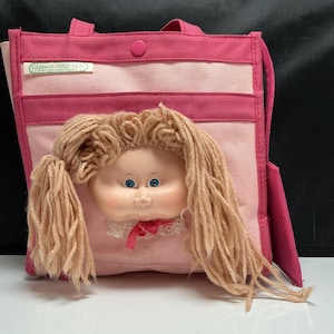 May include: A pink canvas tote bag featuring a Cabbage Patch Kids doll face with yarn hair and blue eyes attached to the front. The bag has a top snap closure, a rectangular pocket, and a white fabric label with green text reading Cabbage Patch Kids.