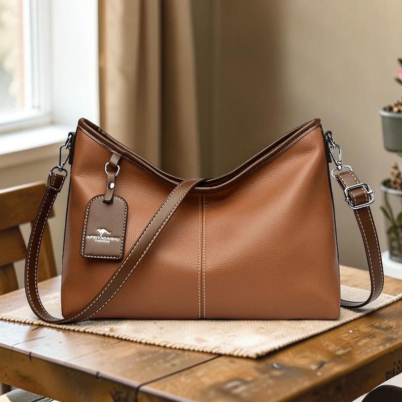 Pu&ograve; includere: Borsa a tracolla in pelle marrone con una lunga tracolla regolabile e un manico pi&ugrave; piccolo. La borsa ha una forma rettangolare con la parte superiore curva e un piccolo tag con testo. La borsa &egrave; appoggiata su un tavolo di legno.