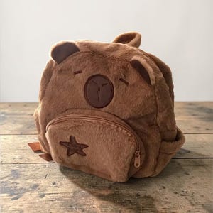 Puede incluir: Una mochila de peluche marrón con forma de oso, con orejas de oso, una cara y un parche en forma de estrella en el bolsillo delantero. Tiene un compartimento principal con cremallera y un bolsillo delantero más pequeño. La mochila está hecha de material suave y difuso.