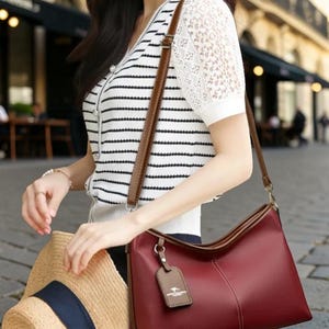 Pu&ograve; includere: Una borsa a tracolla in pelle bordeaux con finiture marroni e un cinturino abbinato. La borsa ha una forma rettangolare e una piccola etichetta attaccata. La persona tiene anche un cappello di paglia con una fascia blu navy.