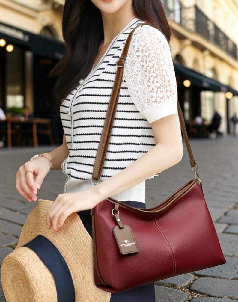 Pu&ograve; includere: Una borsa a tracolla in pelle bordeaux con finiture marroni e un cinturino abbinato. La borsa ha una forma rettangolare e una piccola etichetta attaccata. La persona tiene anche un cappello di paglia con una fascia blu navy.