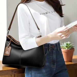 Pu&ograve; includere: Borsa a tracolla nera con finiture e cinturino marroni. La borsa ha un'etichetta con un logo. Una persona indossa una maglietta bianca a maniche lunghe e jeans blu. La maglietta ha la scritta "WOMAN".