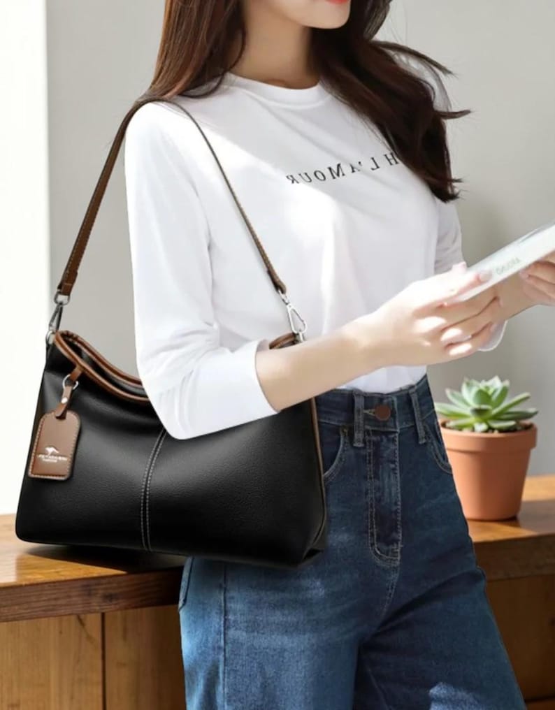 Pu&ograve; includere: Borsa a tracolla nera con finiture e cinturino marroni. La borsa ha un'etichetta con un logo. Una persona indossa una maglietta bianca a maniche lunghe e jeans blu. La maglietta ha la scritta "WOMAN".