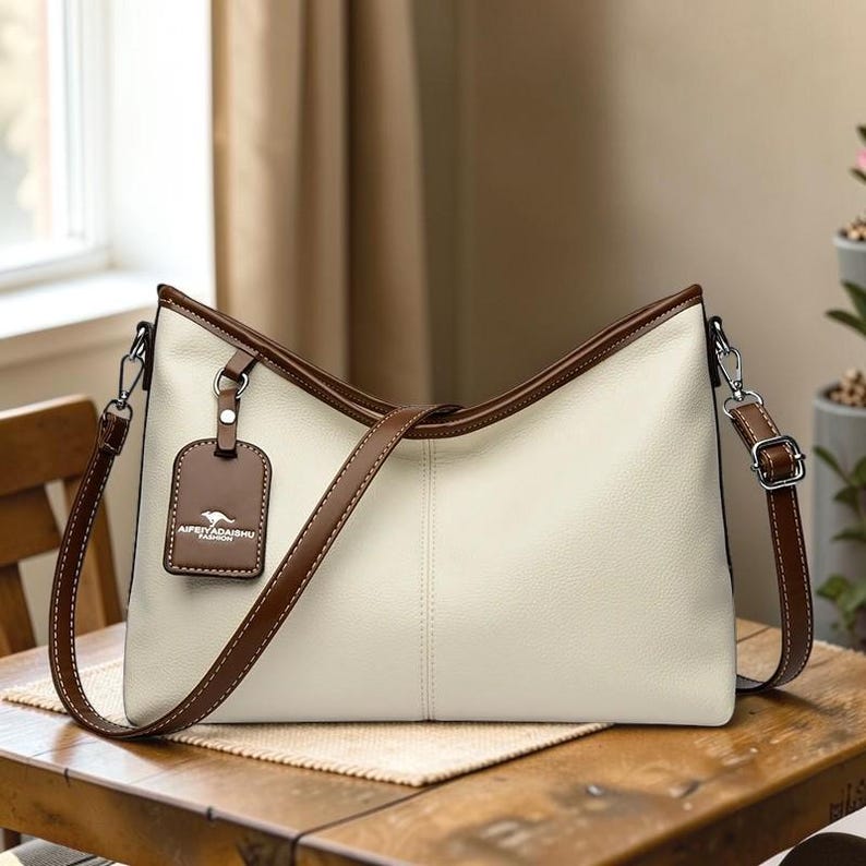 Pu&ograve; includere: Borsa a tracolla color crema con finiture marroni e cinturino abbinato. La borsa presenta un'etichetta rettangolare marrone con la scritta "AIFEYADABSHU". La borsa &egrave; su un tavolo di legno.