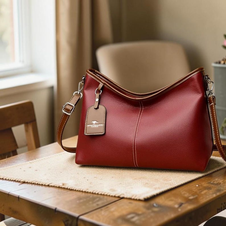 Pu&ograve; includere: Una borsa a tracolla in pelle rossa con finiture marroni e una tracolla regolabile abbinata. La borsa presenta un design cucito e una piccola etichetta con un logo. La borsa &egrave; appoggiata su un tavolo di legno.