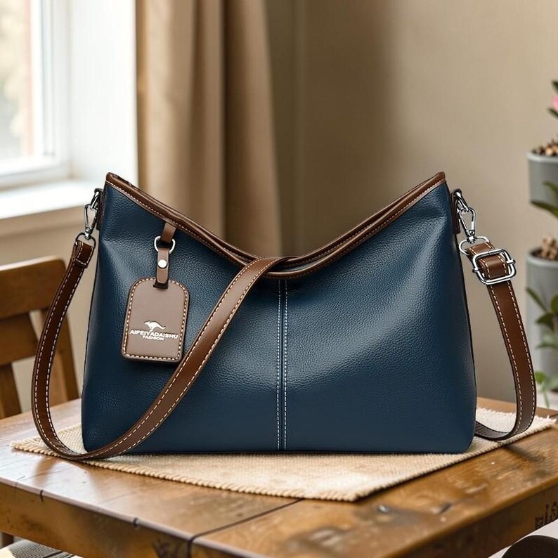 Pu&ograve; includere: Una borsa blu navy con finiture marroni e una tracolla abbinata. La borsa presenta un'etichetta con testo e una cucitura bianca al centro. La borsa &egrave; appoggiata su un tavolo di legno.