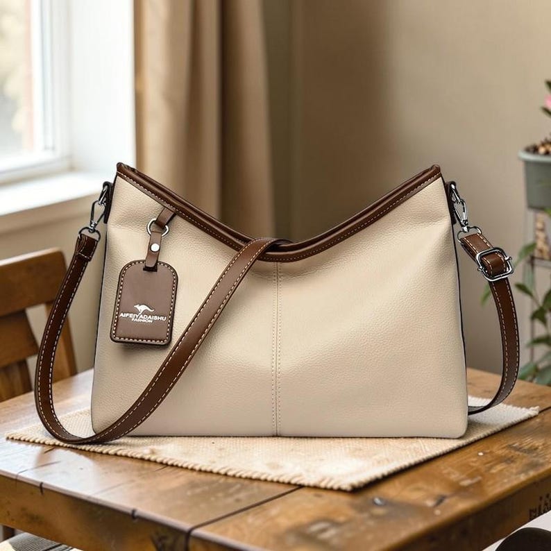Pu&ograve; includere: Borsa a tracolla color crema con finiture marroni e cinturino abbinato. La borsa ha una forma rettangolare con un'etichetta che dice "APENYADAISHU FASHION". La borsa &egrave; su un tavolo di legno.