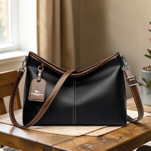 Pu&ograve; includere: Borsa a tracolla nera con finiture e cinturino marroni. La borsa ha una forma rettangolare con la parte superiore curva e una cucitura verticale bianca. Una piccola etichetta pende dalla maniglia.