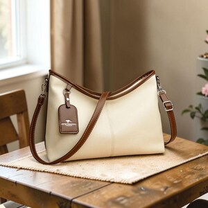 Pu&ograve; includere: Borsa a mano color crema con finiture marroni e una tracolla abbinata. La borsa presenta un'etichetta rettangolare con un logo. La borsa &egrave; su un tavolo di legno.