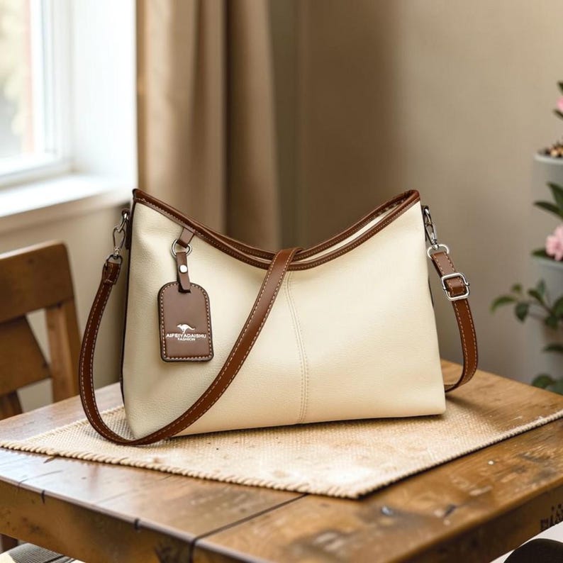 Pu&ograve; includere: Borsa a mano color crema con finiture marroni e una tracolla abbinata. La borsa presenta un'etichetta rettangolare con un logo. La borsa &egrave; su un tavolo di legno.
