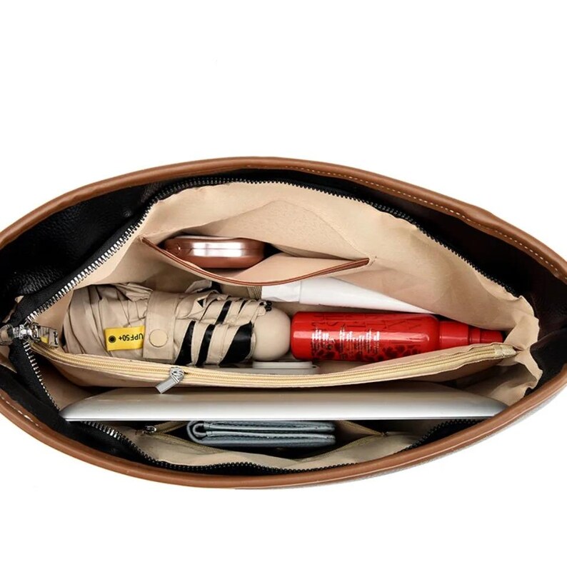 Pu&ograve; includere: Vista aperta di una borsa in pelle nera e marrone piena di oggetti essenziali. All'interno, un interno beige contiene un compatto, un piccolo ombrello, una bottiglia rossa, un tablet e un portafoglio. La borsa ha una chiusura con cerniera e un bordo marrone.