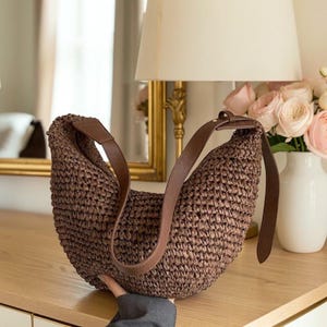 May include: A brown woven shoulder bag with a brown leather strap. The bag has a curved shape and a textured surface. The strap is attached to the bag with a small leather piece. The bag is on a wooden surface.