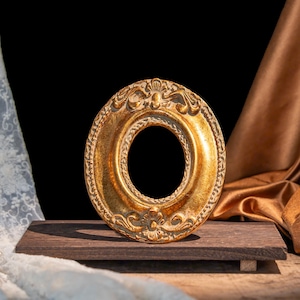 Könnte beinhalten: Ein ovaler Bilderrahmen mit metallisch goldener Oberfläche und detaillierten Prägungen entlang des Randes. Der Rahmen verfügt über eine zentrale Öffnung und steht aufrecht auf einem dunklen, rechteckigen Holzsockel mit strukturierter Oberfläche.