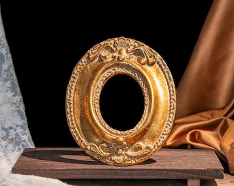 May include: An oval-shaped picture frame with a metallic gold finish and intricate embossed detailing along the border. The frame features a central opening and is positioned upright on a dark, rectangular wooden base, showcasing a textured surface.
