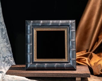 May include: A square picture frame with a dark, metallic, bamboo-textured border and an inner gold-toned decorative trim. The frame is positioned on a dark wooden rectangular base, showcasing a deep black centre area within the frame's structure.