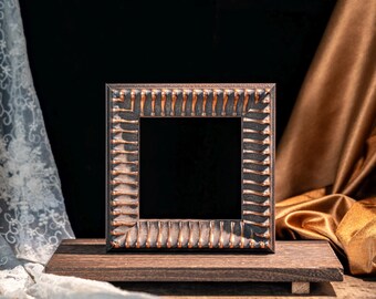 May include: A square picture frame with a dark, textured finish and copper-coloured highlights along its ridged edges. The frame is mounted on a rectangular wooden base, creating a stable display piece with a distinct contrast between the dark frame and metallic accents.