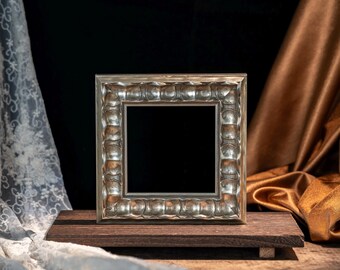 May include: A square picture frame with a metallic silver finish and a repeating scalloped, rounded edge pattern. The frame features a smooth inner border and a deep, dark center opening, presented as a standalone decorative object.