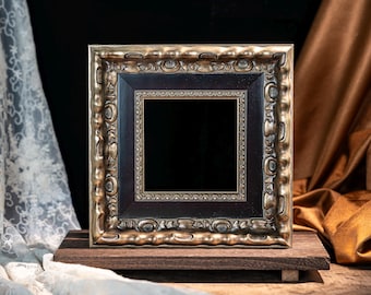May include: A square picture frame featuring an ornate, metallic gold outer border with a dark, smooth inner frame and a decorative, textured gold trim surrounding the central black opening, all resting on a dark wooden rectangular base.
