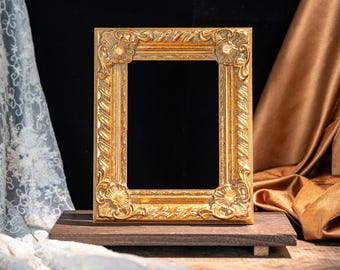 May include: A rectangular picture frame with a metallic gold finish, featuring ornate relief carvings of floral motifs at each corner and a textured, rope-like pattern along the inner and outer borders, standing upright on a dark wooden base.