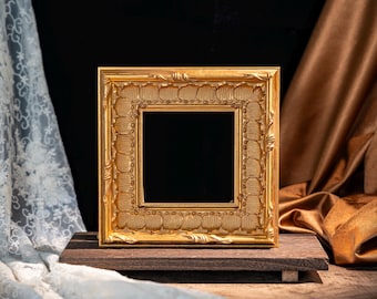 May include: A square picture frame with a metallic gold finish, featuring an ornate, textured border with repeating leaf-like patterns and raised decorative elements along the edges, surrounding a deep, empty, dark-coloured centre opening.