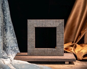 May include: A square picture frame with a dark, textured, bronze-toned finish featuring vertical ridges. The frame is mounted on a rectangular, dark brown wooden base with small feet, creating a stable display stand for a photograph or artwork.