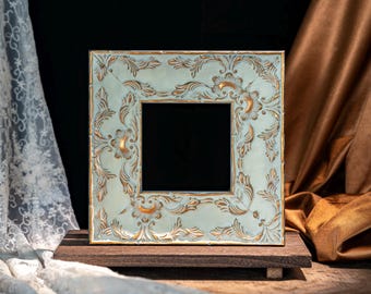 May include: A square picture frame featuring a pale mint green finish with intricate, raised gold-toned floral and scrollwork relief patterns. The frame has a central square opening and is positioned on a dark brown rectangular wooden base.