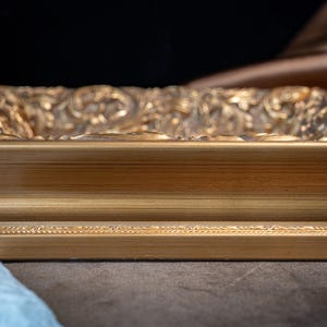 May include: A rectangular gold picture frame featuring a smooth, flat inner border and a highly detailed, ornate outer edge with intricate scrollwork and relief patterns. The frame has a metallic finish with subtle highlights and deep shadows.