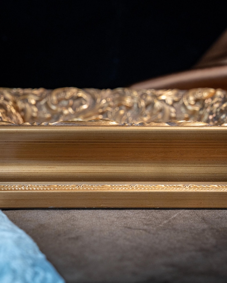 May include: A rectangular gold picture frame featuring a smooth, flat inner border and a highly detailed, ornate outer edge with intricate scrollwork and relief patterns. The frame has a metallic finish with subtle highlights and deep shadows.