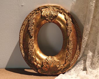 May include: An oval-shaped picture frame with a metallic gold finish and a distressed texture. The frame features raised relief carvings of leaves and floral garlands along its border, creating a textured appearance around the central opening.