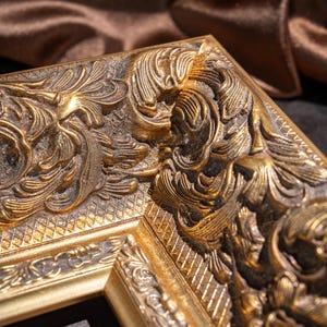 May include: A corner section of a rectangular picture frame featuring an ornate, raised relief pattern with gold-toned metallic finish and dark, recessed detailing. The frame displays intricate scrollwork and a cross-hatched texture.