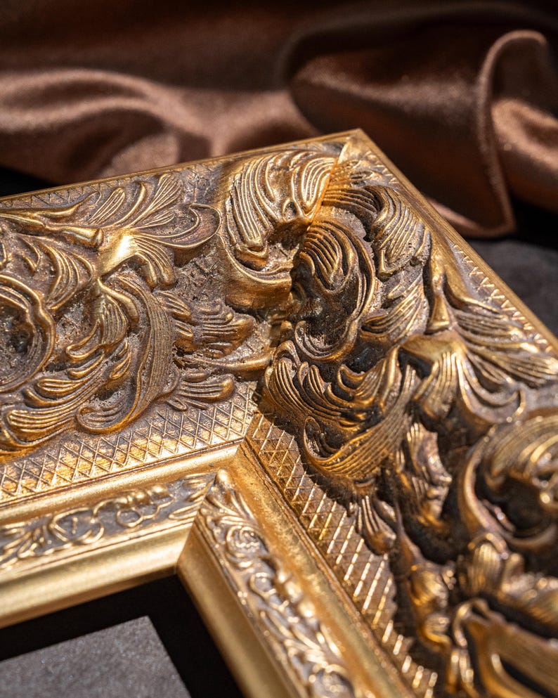 May include: A corner section of a rectangular picture frame featuring an ornate, raised relief pattern with gold-toned metallic finish and dark, recessed detailing. The frame displays intricate scrollwork and a cross-hatched texture.