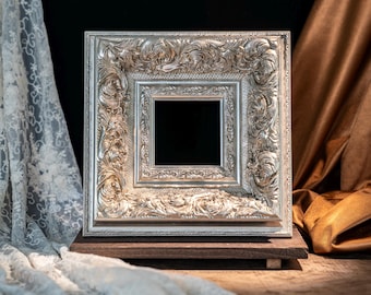 May include: A square picture frame with a metallic silver finish and intricate, raised scrollwork patterns. The frame features a deep, multi-layered border design with a central square opening, resting on a dark brown wooden base.