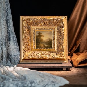 May include: A square landscape painting featuring a river, trees, and distant mountains, encased in a wide, ornate, gold-colored frame with intricate relief carvings. The artwork is displayed on a dark wooden base, positioned between draped fabrics.
