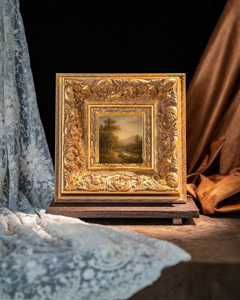 May include: A square landscape painting featuring a river, trees, and distant mountains, encased in a wide, ornate, gold-colored frame with intricate relief carvings. The artwork is displayed on a dark wooden base, positioned between draped fabrics.