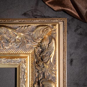May include: A rectangular picture frame with a metallic gold finish and dark brown antiquing in the recessed areas. The frame features a wide, ornate border with raised relief scrollwork, acanthus leaf motifs, and a cross-hatched texture.