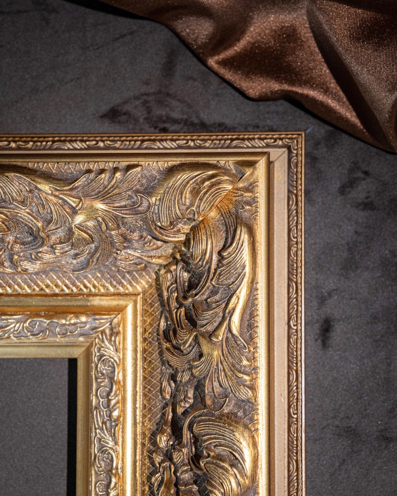 May include: A rectangular picture frame with a metallic gold finish and dark brown antiquing in the recessed areas. The frame features a wide, ornate border with raised relief scrollwork, acanthus leaf motifs, and a cross-hatched texture.