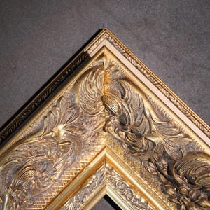 May include: A gold-colored picture frame corner featuring intricate, raised relief scrollwork and floral patterns. The frame has a metallic finish with dark, recessed areas that highlight the detailed carvings along the outer and inner edges.
