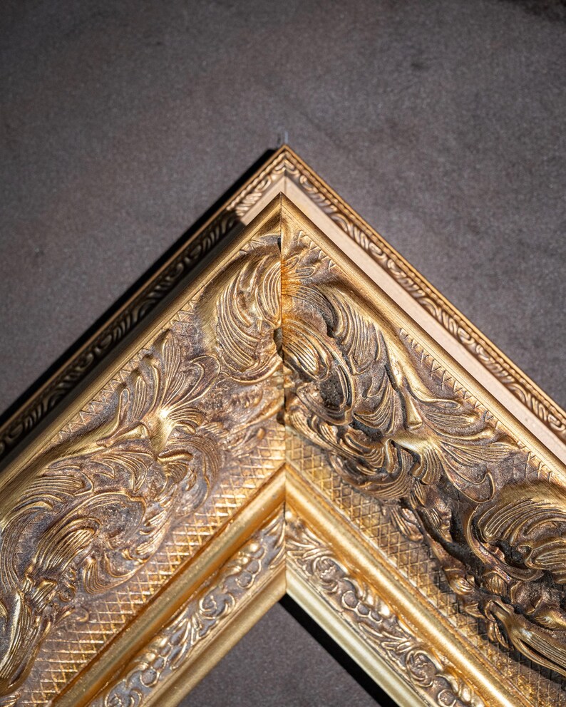 May include: A gold-colored picture frame corner featuring intricate, raised relief scrollwork and floral patterns. The frame has a metallic finish with dark, recessed areas that highlight the detailed carvings along the outer and inner edges.