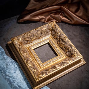 May include: A square picture frame with a metallic gold finish, featuring intricate raised scrollwork and floral relief patterns along the wide border. The frame has a stepped profile with a smooth inner edge and a textured, recessed section.