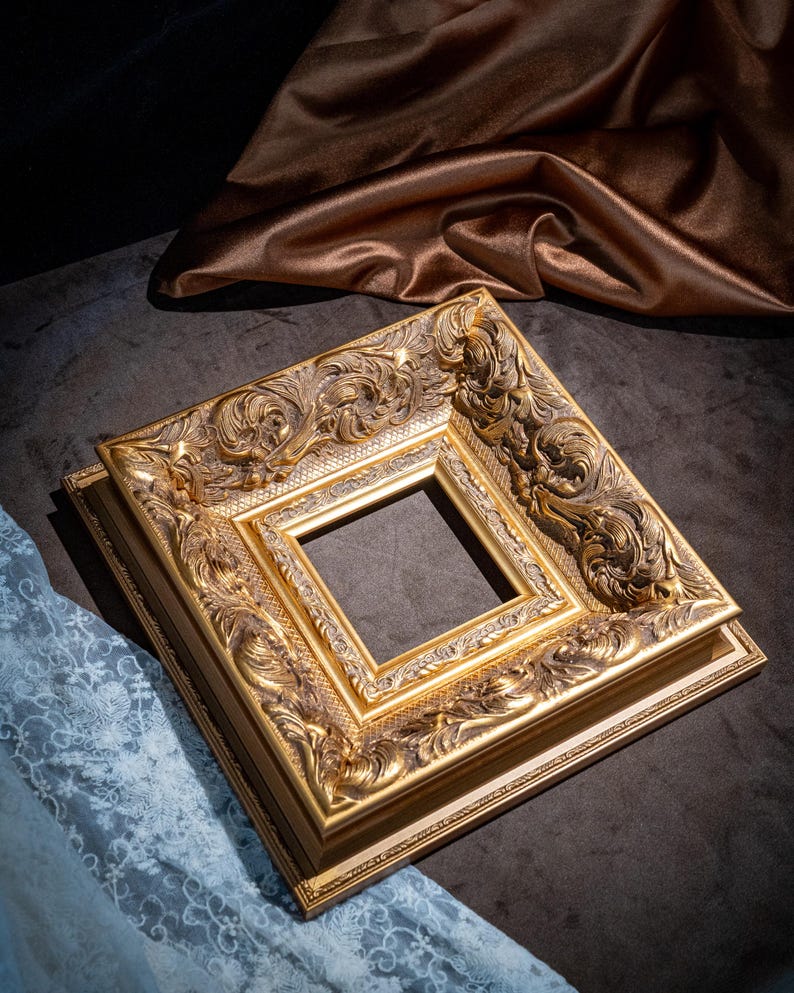 May include: A square picture frame with a metallic gold finish, featuring intricate raised scrollwork and floral relief patterns along the wide border. The frame has a stepped profile with a smooth inner edge and a textured, recessed section.