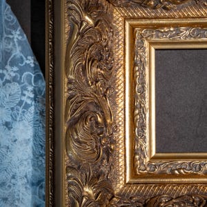 May include: A rectangular picture frame with a metallic gold finish and a dark inner border. The frame features intricate, raised relief carvings of scrolling foliage and leaves, complemented by a textured cross-hatched pattern along the inner edge.