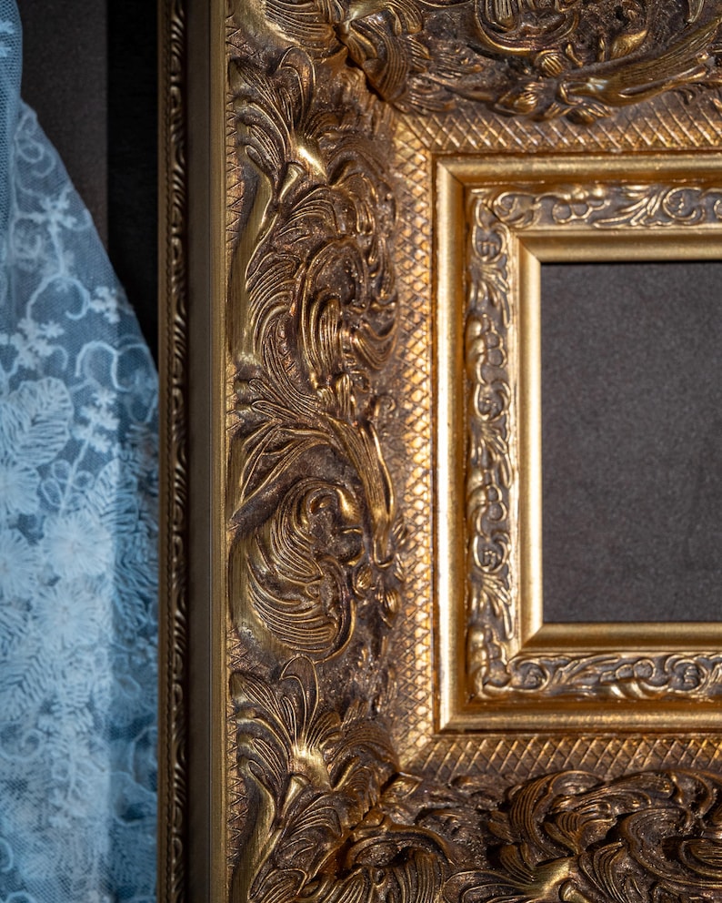 May include: A rectangular picture frame with a metallic gold finish and a dark inner border. The frame features intricate, raised relief carvings of scrolling foliage and leaves, complemented by a textured cross-hatched pattern along the inner edge.