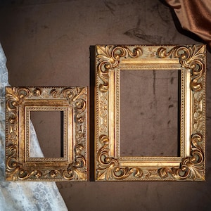 May include: Two rectangular picture frames with ornate, raised scrollwork and leaf motifs. The frames feature a metallic gold finish with distressed, muted green undertones, showcasing a beaded inner border and a wide, decorative outer edge.