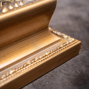 May include: A gold-colored picture frame corner featuring a stepped, concave profile and a decorative beaded edge detail. The frame has a brushed metallic finish with visible horizontal graining and a raised relief pattern along the top edge.