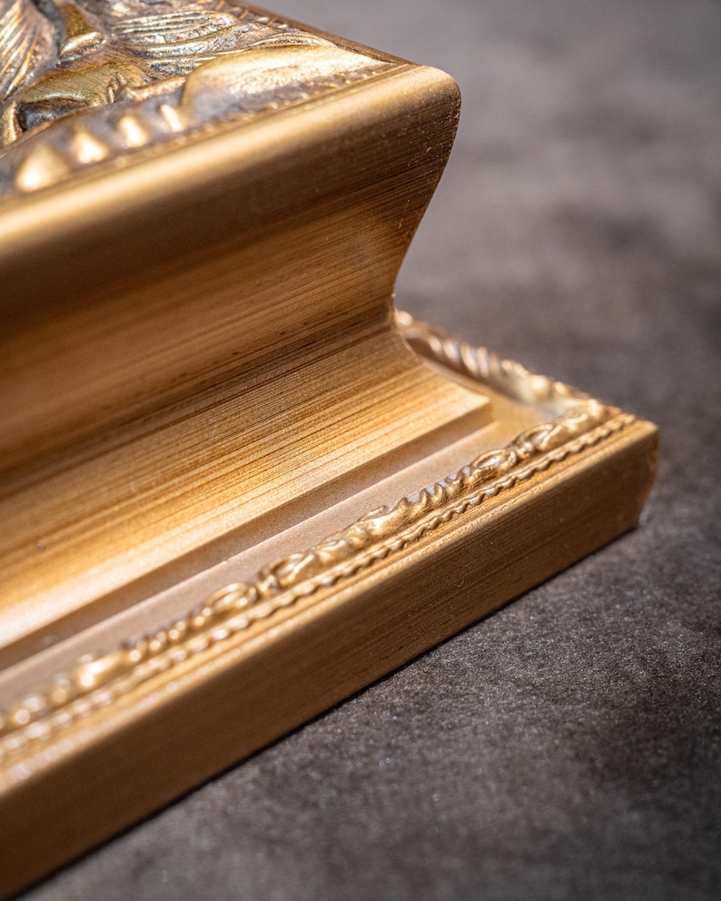 May include: A gold-colored picture frame corner featuring a stepped, concave profile and a decorative beaded edge detail. The frame has a brushed metallic finish with visible horizontal graining and a raised relief pattern along the top edge.