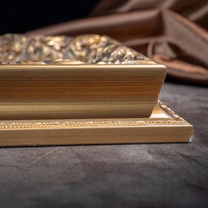 May include: A rectangular picture frame with a metallic gold finish. The frame features a stepped profile with a smooth, flat inner edge and a decorative, ornate outer border with intricate relief detailing along the top and side edges.