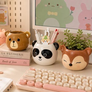 Op de afbeelding: Een verzameling decoratieve keramische containers in de vorm van een beer, panda en hert. De beer is bruin, de panda is zwart en wit en het hert is bruin en bruingeel. De containers staan op een bureau met een toetsenbord en boeken.