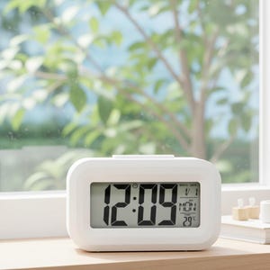 Può includere: Una sveglia digitale bianca che mostra l'ora 12:09, la data e la temperatura. L'orologio ha una forma rettangolare con angoli arrotondati. È su una superficie di legno, con una finestra e fogliame verde sullo sfondo.